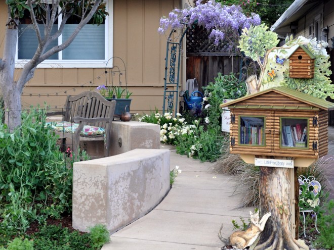 A charming garden scene featuring a stone pathway, a wooden bench with colorful cushions, a whimsical little free library made from wood, and vibrant greenery, including wisteria flowers hanging above.