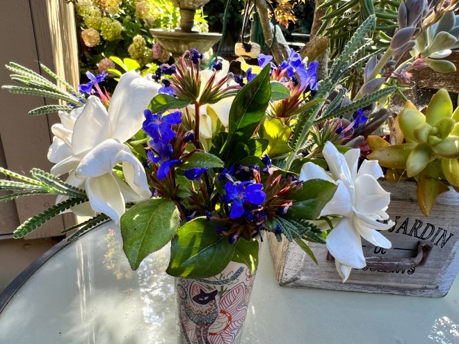 A colorful floral arrangement featuring white gardenias and vibrant blue flowers, displayed in a decorative cup on a table.