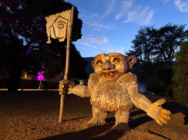 A wooden troll sculpture holding a flag, set in a garden during twilight with trees in the background.
