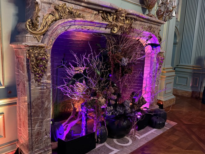 A beautifully decorated fireplace featuring Halloween-themed decor, including purple lighting, black crows, and ghostly figures, surrounded by dried branches and flowers.
