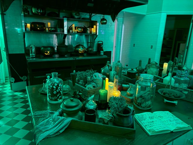 A dimly lit kitchen with green lighting, featuring a variety of jars, herbs, and candles on a table, along with Halloween-themed decorations and carved pumpkins displayed on shelves.