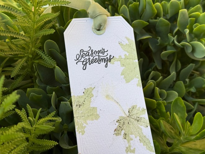 A gift tag with the text 'Season's Greetings' surrounded by green leaf impressions, resting among lush green foliage.