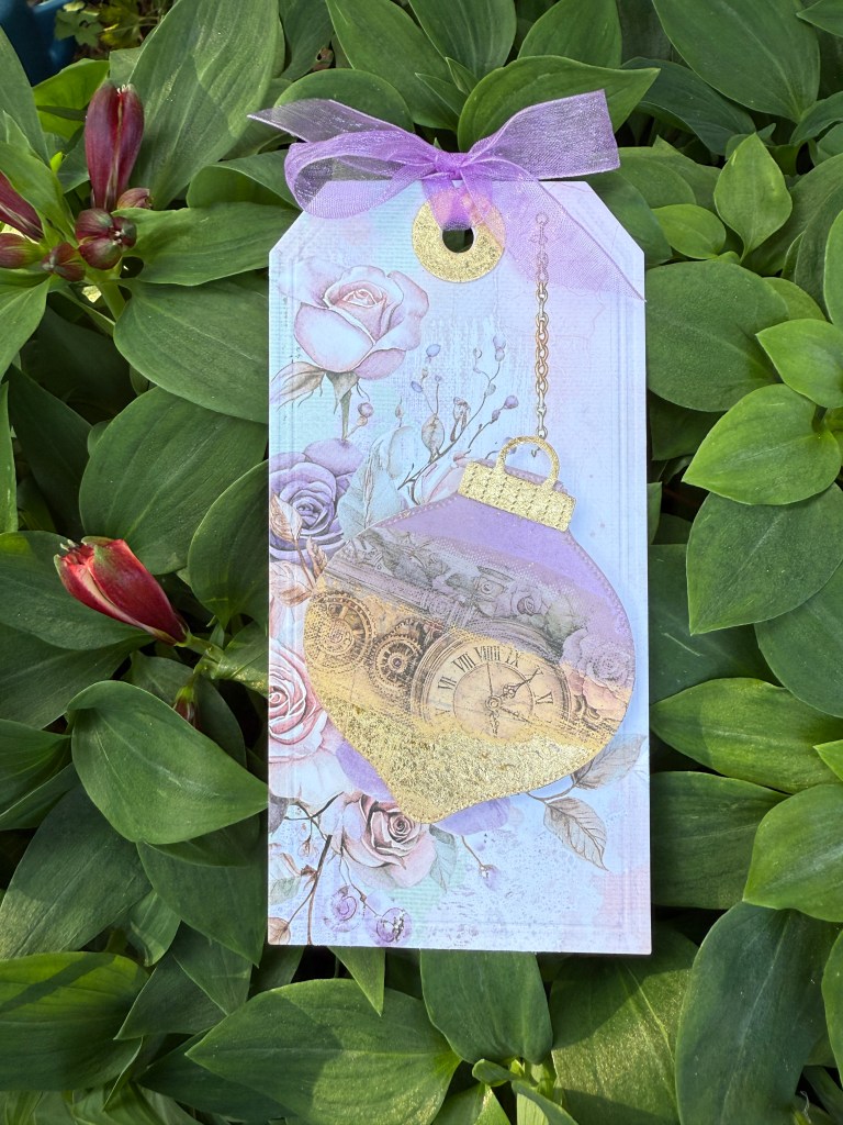 A decorative gift tag featuring a clock motif surrounded by rose illustrations, with a purple ribbon and gold accents, set against lush green leaves.