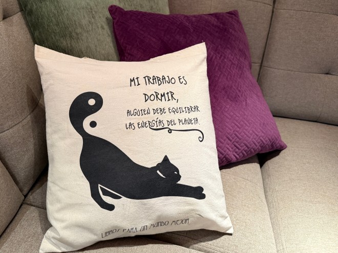 A decorative cushion featuring a black cat silhouette and a humorous Spanish text that translates to 'My job is to sleep, someone must balance the energies of the planet.' The cushion is sitting on a gray couch next to a purple pillow.