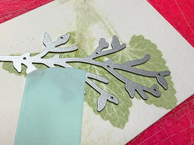 A metallic silver leaf branch cutout positioned on a textured green leaf background, with a piece of light blue paper partially covering it.