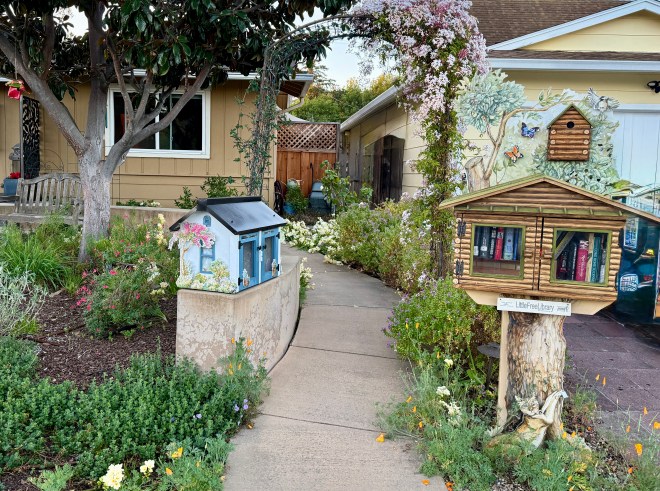A charming garden pathway lined with colorful flowers leads to two unique book-sharing structures—the left one is a small blue library box, and the right is a wooden book house decorated with a birdhouse, surrounded by lush greenery.