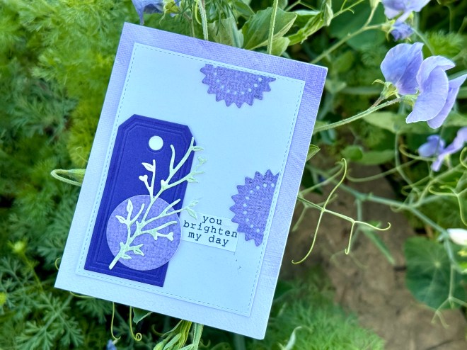 A handmade greeting card featuring a purple tag and floral design with the text 'you brighten my day' set against a background of green foliage and purple flowers.
