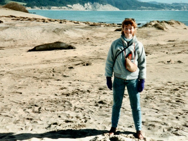 A woman wearing a gray sweater and blue jeans stands on a sandy beach with a seal resting in the background, overlooking a serene ocean view.