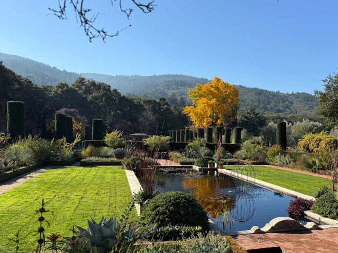 A serene garden landscape featuring well-manicured lawns, a reflecting pond, and neatly trimmed hedges. In the background, hills are visible under a clear blue sky, with a prominent yellow tree adding a splash of color.
