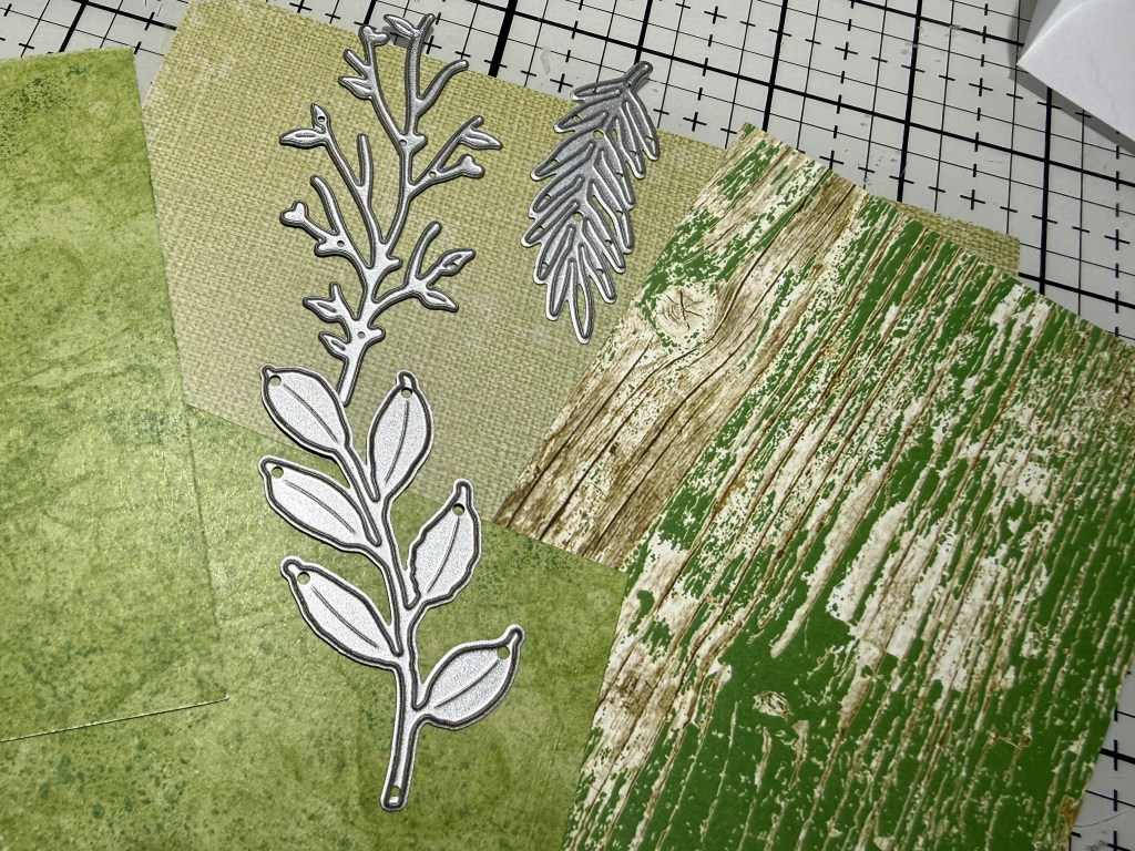 Various pieces of textured green craft paper and metal leaf die cuts arranged on a grid cutting mat.