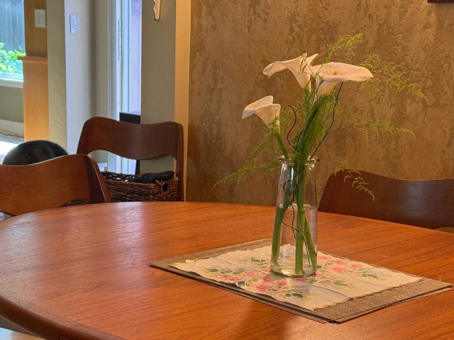 A wooden dining table set with a floral centerpiece featuring white calla lilies in a glass vase. The table is adorned with a decorative cloth, and wooden chairs are positioned around it.