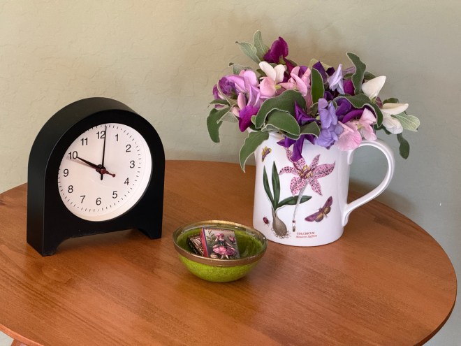 A black clock displaying the time on a wooden table next to a white pitcher filled with vibrant purple and pink flowers, accompanied by a small green bowl containing decorative cards.