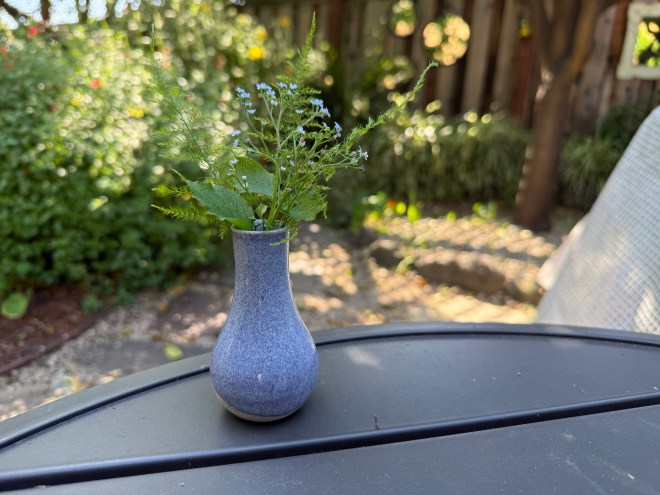 A small blue vase holding a few green leaves and delicate blue flowers, placed on a black table in a garden setting with blurred greenery in the background.