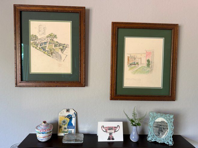 Two framed illustrations hanging on a wall, featuring architectural sketches. Below them is a decorative table with various items, including a colorful pot, a small painting, and a vase with greenery.