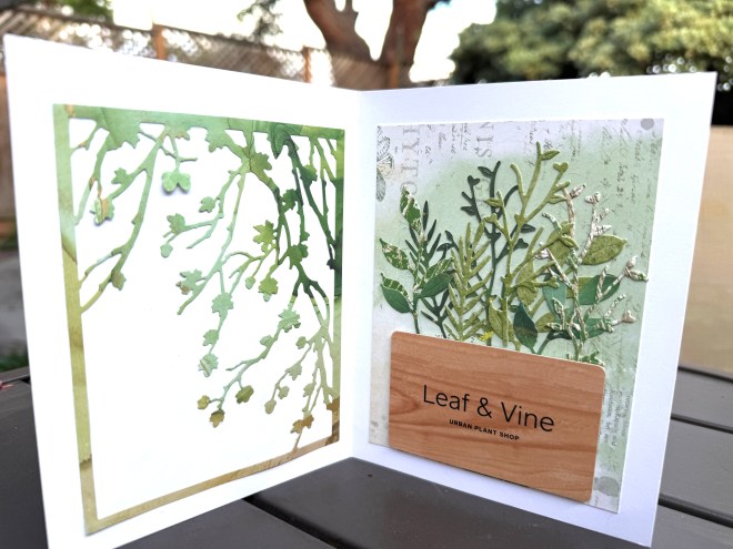 An open greeting card featuring botanical illustrations, with a cut-out design on the left and a layered green foliage composition on the right, along with a wooden label for 'Leaf & Vine Urban Plant Shop'.