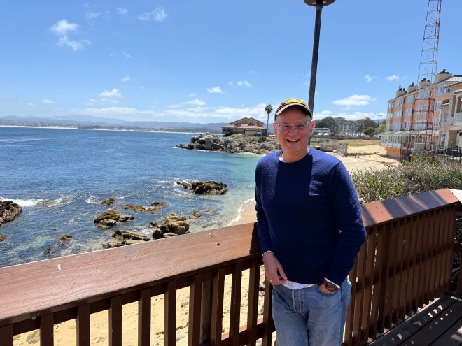 A man smiling while standing on a wooden deck by the shore, with rocky coastline and blue ocean in the background under a clear sky.
