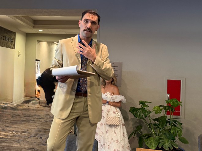 A man in a tan suit and patterned tie stands holding a rolled document, looking thoughtfully at someone. A woman in a white dress stands in the background, partially obscured by a plant.