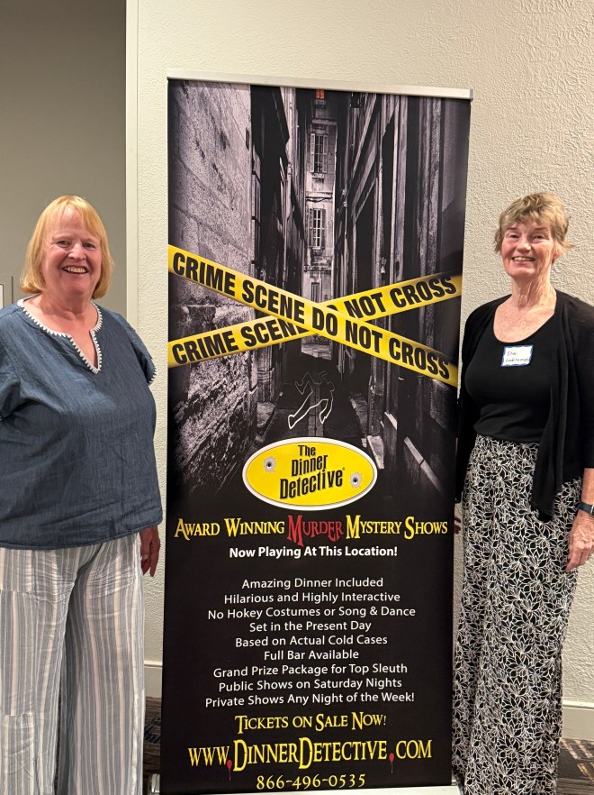 Two women standing next to a banner for 'The Dinner Detective,' featuring a crime scene motif and promoting murder mystery shows.