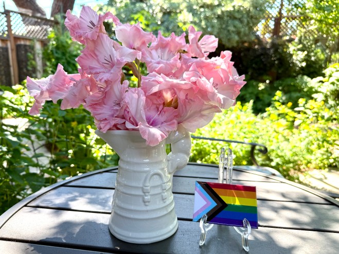 A white vase filled with pink flowers, placed on a table outdoors, beside a small stand displaying a rainbow pride flag card.