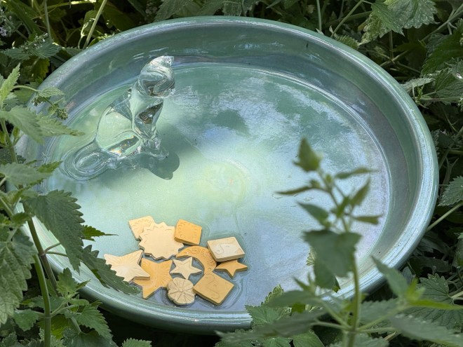 A shallow green birdbath surrounded by greenery, featuring a water fountain shaped like a dinosaur and a collection of yellow, star-shaped toys at the bottom.
