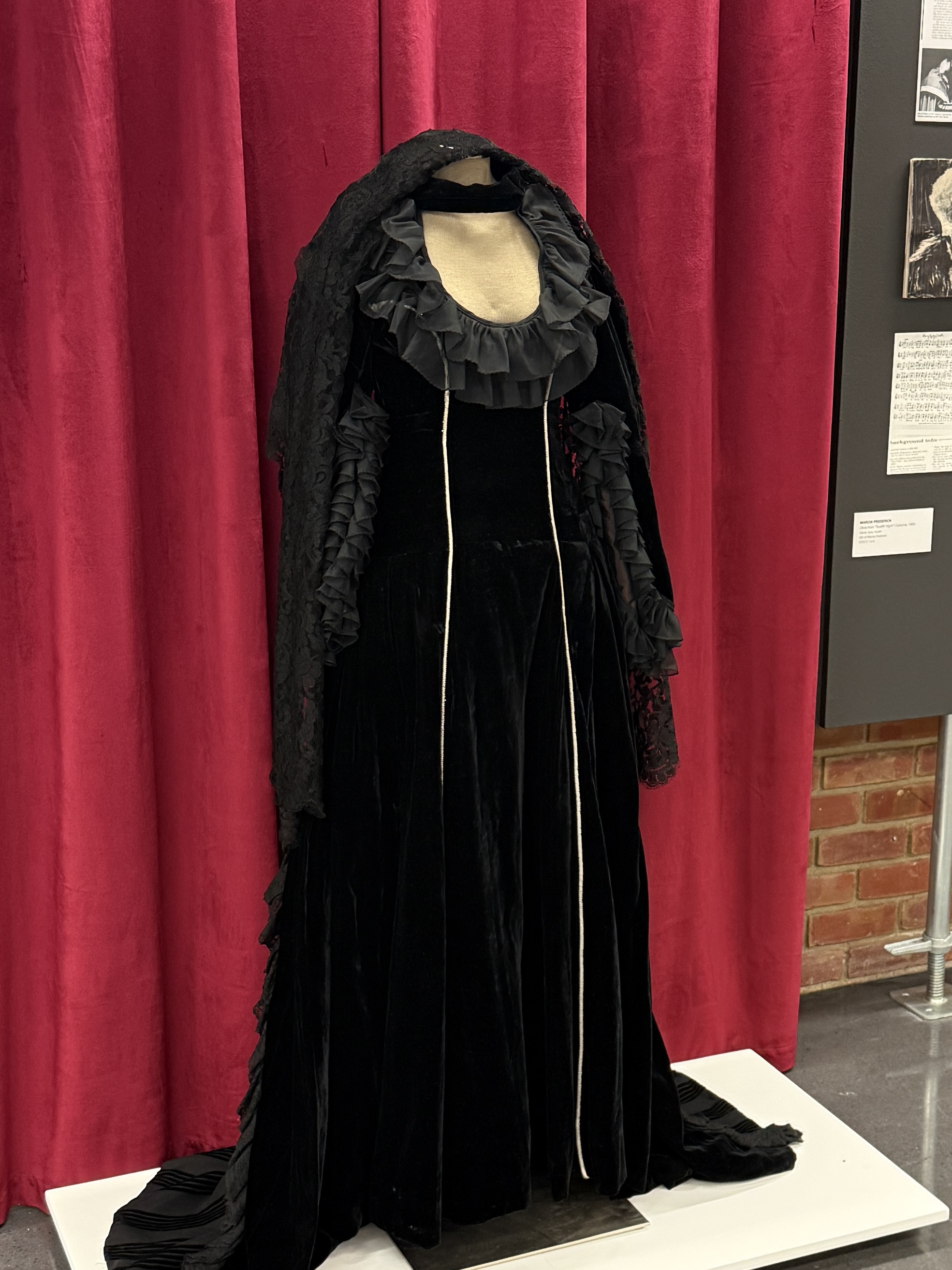 A black velvet gown and accompanying black lace mantilla. The black dress is largely plain, with black ruffles at the sleeve openings and along the neckline, with two bands of silver piping down the front. On the back of the dress' skirt, there is black striped detailing, with additional black lace trim. The dress opens in the back with a silver zipper. The black mantilla features a floral pattern, and has wavy edges.