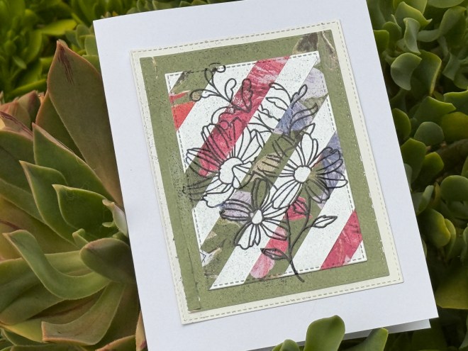 A handmade greeting card featuring a floral design outlined in black, layered over colorful diagonal stripes on a textured background, set against a backdrop of succulent plants.