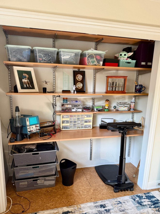 A neatly organized closet workspace featuring shelves with storage bins, tools, and decorative items. The setup includes a standing desk, smaller containers for supplies, a framed photo, and a few mechanical devices.