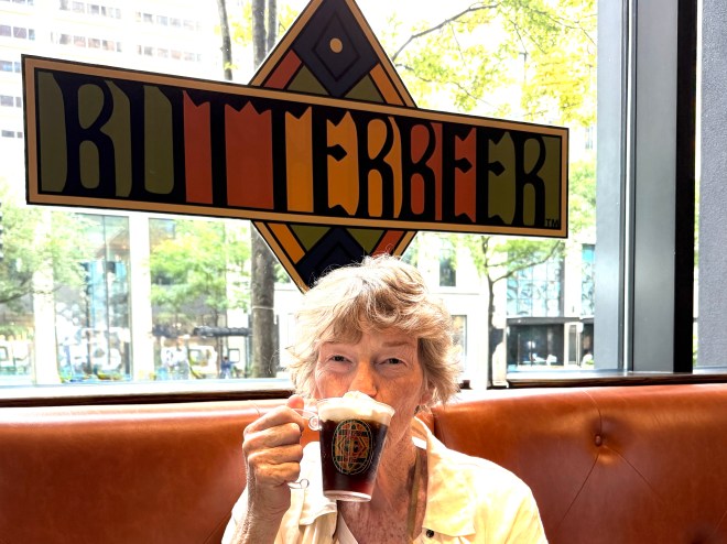 A person enjoying Butterbeer in a cafe with a window view, smiling while holding a cup with foam on top, under a colorful 'Butterbeer' sign.