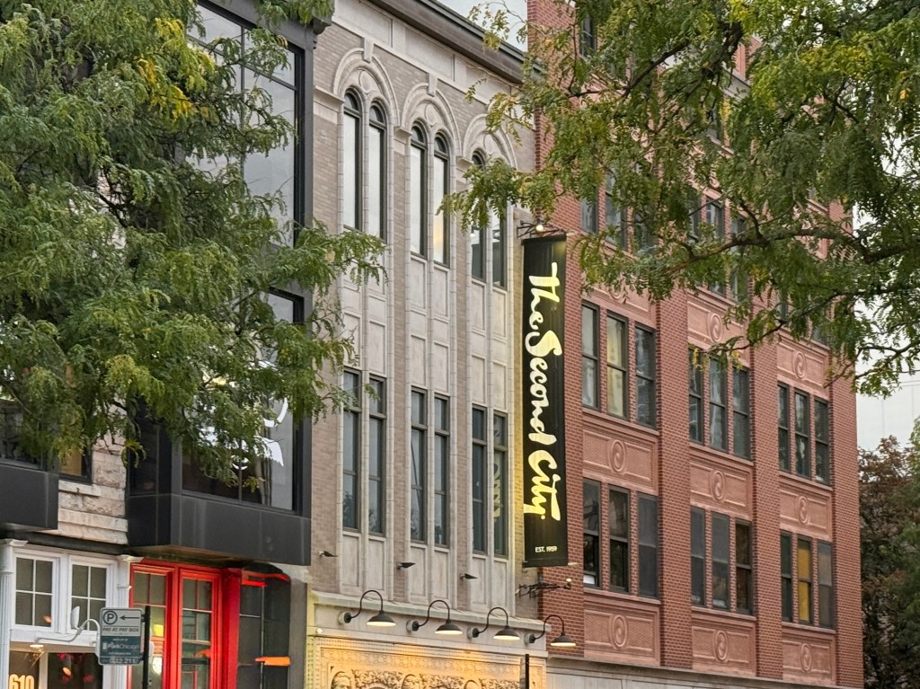Exterior view of The Second City building featuring a prominent yellow sign and decorative architecture, surrounded by trees and urban elements.