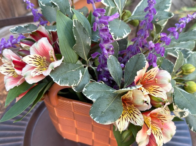 A decorative basket filled with colorful flowers, including pink and orange lilies and purple flowering plants, surrounded by green foliage.