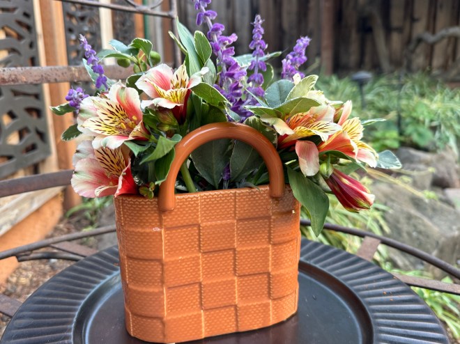 A small, woven-style orange basket filled with colorful flowers, including purple and pink blooms, sitting on a decorative black plate outdoors.