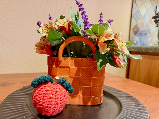 A woven basket filled with colorful flowers, including purple and orange blooms, placed on a wooden table next to a crocheted red tomato with green leaves.